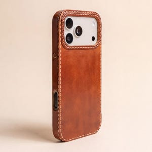 Puede incluir: Una funda de cuero marrón para teléfono con un acabado mate suave y costuras visibles de color claro a lo largo de los bordes. La funda incluye un recorte rectangular para el módulo de la cámara, que muestra tres lentes circulares y un flash.
