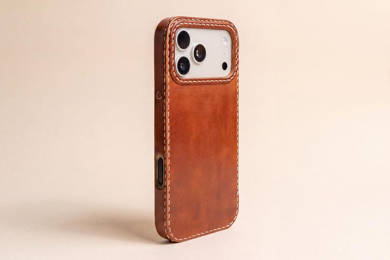 May include: A brown leather phone case featuring a smooth, matte finish and visible light-colored stitching along the edges. The case includes a rectangular cutout for the camera module, which reveals three circular lenses and a flash component.