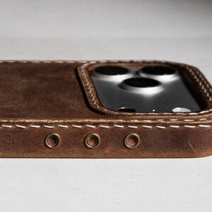 May include: A brown leather phone case featuring visible white stitching along the edges and three small circular metal eyelets on the side. The case has a raised cutout section surrounding the triple camera lens array of the smartphone.