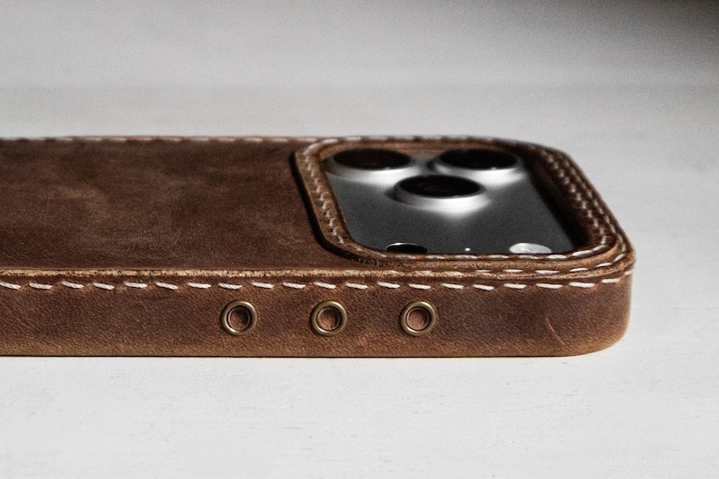 May include: A brown leather phone case featuring visible white stitching along the edges and three small circular metal eyelets on the side. The case has a raised cutout section surrounding the triple camera lens array of the smartphone.