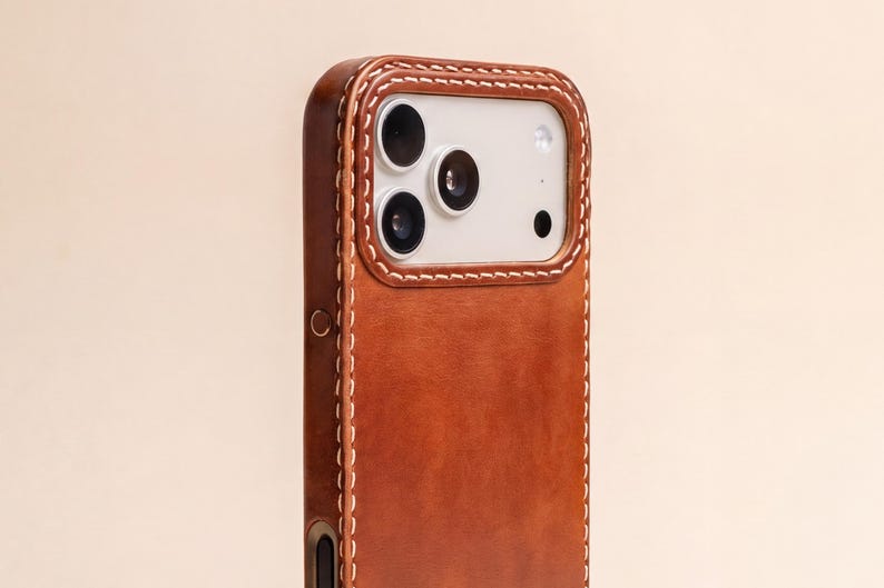 May include: A tan leather phone case featuring a smooth, matte finish and prominent white contrast stitching along the edges. The case includes a raised, rectangular cutout section that frames the three camera lenses of the smartphone.
