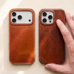 May include: Two rectangular phone cases crafted from smooth, tan-colored leather with visible light-colored stitching along the edges. Each case features a cutout for the camera module, exposing the white back of the phone and the lens array.