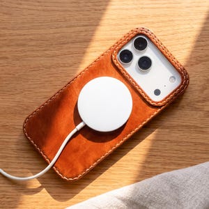 May include: A tan leather smartphone case with visible stitching around the edges, featuring a circular white magnetic wireless charger attached to the back. The case surrounds a smartphone with a triple-lens camera system and a white back panel.