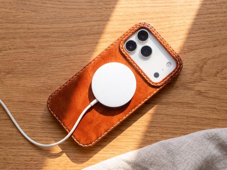 May include: A tan leather smartphone case with visible stitching around the edges, featuring a circular white magnetic wireless charger attached to the back. The case surrounds a smartphone with a triple-lens camera system and a white back panel.
