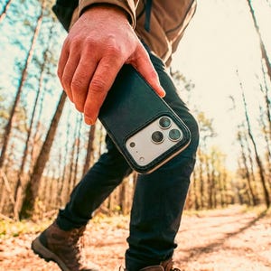 May include: A dark green leather smartphone case with a visible camera cutout featuring three circular lenses. The case has a smooth texture and stitched edges, designed to fit a modern smartphone with a prominent rear camera array system.