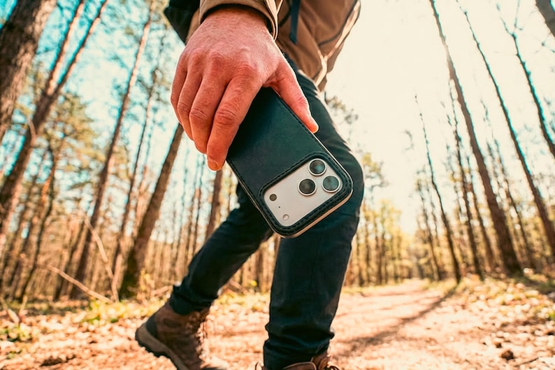 May include: A dark green leather smartphone case with a visible camera cutout featuring three circular lenses. The case has a smooth texture and stitched edges, designed to fit a modern smartphone with a prominent rear camera array system.