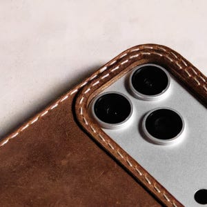 May include: A brown leather phone case featuring visible white stitching around the camera cutout. The case surrounds a silver-toned smartphone with three circular camera lenses and a flash, showcasing a textured finish on the leather surface.