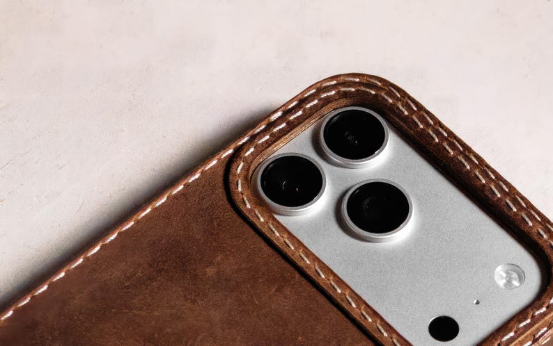 May include: A brown leather phone case featuring visible white stitching around the camera cutout. The case surrounds a silver-toned smartphone with three circular camera lenses and a flash, showcasing a textured finish on the leather surface.