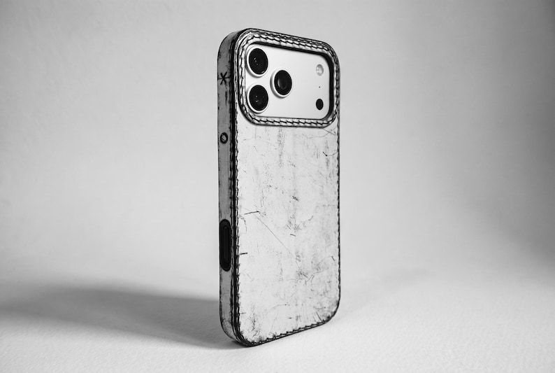 May include: A black and white phone case with a distressed, weathered texture and visible stitching along the edges. The case features a cutout for the camera module, which contains three circular lenses, and a small star symbol on the side.