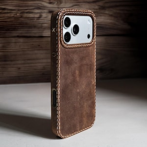 May include: A brown leather phone case with visible light-colored stitching along the edges. The case features a cutout for the camera module, which contains three circular lenses and a flash. A small white X is stitched near the top left corner.