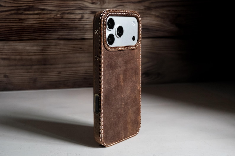 May include: A brown leather phone case with visible light-colored stitching along the edges. The case features a cutout for the camera module, which contains three circular lenses and a flash. A small white X is stitched near the top left corner.