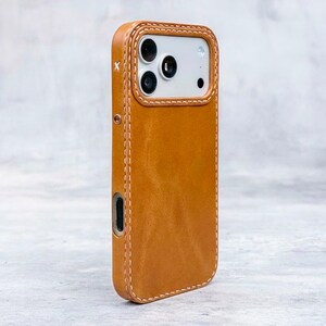 May include: A tan leather phone case featuring visible white stitching along the edges and a cutout for the camera lenses. The case has a small white letter X printed on the side near the top and a metallic button detail on the side frame.