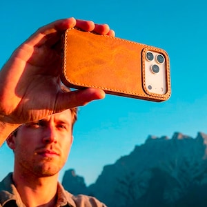 May include: A tan leather phone case with visible stitching along the edges, featuring a rectangular cutout for the camera lenses. The case is designed to fit a smartphone, showing the back panel with a smooth, matte texture and a raised border.