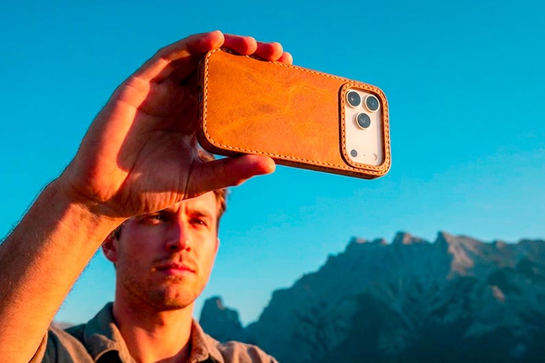 May include: A tan leather phone case with visible stitching along the edges, featuring a rectangular cutout for the camera lenses. The case is designed to fit a smartphone, showing the back panel with a smooth, matte texture and a raised border.