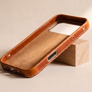 May include: A tan leather phone case with visible white stitching along the edges. The interior features a soft, suede-like lining, and the exterior includes precise cutouts for the camera module and side buttons, showcasing a natural grain texture.