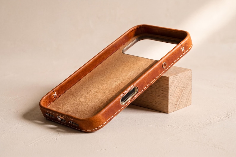 May include: A tan leather phone case with visible white stitching along the edges. The interior features a soft, suede-like lining, and the exterior includes precise cutouts for the camera module and side buttons, showcasing a natural grain texture.