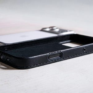 May include: A black leather phone case with visible stitching along the edges, featuring precise cutouts for side buttons and ports, positioned in front of a white smartphone with a metallic camera module on the back.