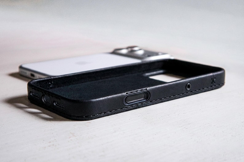 May include: A black leather phone case with visible stitching along the edges, featuring precise cutouts for side buttons and ports, positioned in front of a white smartphone with a metallic camera module on the back.