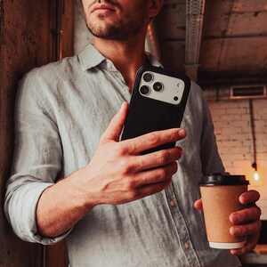 May include: A black smartphone case with a light-colored camera module cutout, held in a hand alongside a brown paper coffee cup. The case features a sleek, solid black back panel and a protective frame, showcasing a modern design for mobile devices.