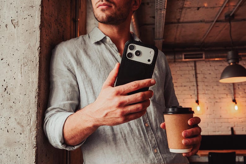 May include: A black smartphone case with a light-colored camera module cutout, held in a hand alongside a brown paper coffee cup. The case features a sleek, solid black back panel and a protective frame, showcasing a modern design for mobile devices.