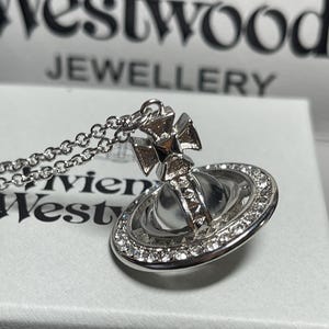 May include: Silver pendant necklace with a planet-shaped charm. The charm has a clear orb, a crystal-studded halo, and a cross-shaped detail. The necklace is on a fine chain. The word "JEWELLERY" is visible in the background.
