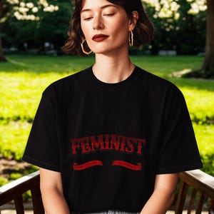 Puede incluir: Camiseta negra con la palabra "FEMINIST" en texto rojo estilizado. La camiseta tiene cuello redondo y mangas cortas. La persona lleva vaqueros azules y pendientes de aro dorados. El fondo es un parque con hierba verde y árboles.