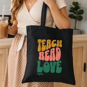 Puede incluir: Bolso tote negro con las palabras "TEACH READ LOVE" en letras de estilo retro. Las palabras son de color amarillo, coral y verde. El nombre "Sophia" está impreso debajo. El bolso lo lleva una persona.