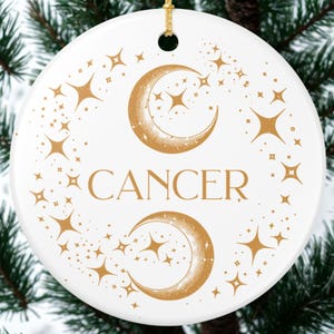 Puede incluir: Un adorno redondo de cerámica blanca con un gráfico de color dorado de dos lunas crecientes y varias formas de estrellas. La palabra CANCER está impresa en letras mayúsculas doradas en el centro. Un cordón dorado está unido arriba.