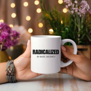 Puede incluir: Taza de cerámica blanca con las palabras "RADICALIZED BY BASIC DECENCY" impresas en negro. La taza está sostenida por dos manos, con un fondo borroso de flores y luces.
