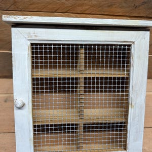 May include: A square wooden cabinet with a distressed white painted finish. The door features a silver metal wire mesh screen and a small round wooden knob. Inside, the cabinet is divided into four equal compartments with natural wood shelves.