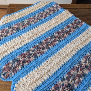 May include: A handmade crochet blanket with a scalloped edge. The blanket features alternating stripes of light blue, cream, and patterned sections with shades of purple and gray. The blanket is made of soft yarn.