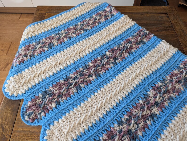 May include: A handmade crochet blanket with a scalloped edge. The blanket features alternating stripes of light blue, cream, and patterned sections with shades of purple and gray. The blanket is made of soft yarn.