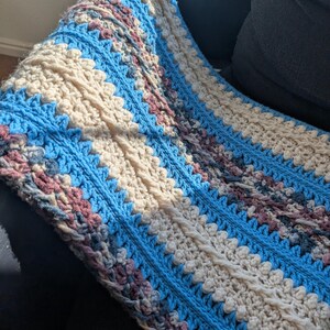 May include: A handmade crochet blanket with a textured design. The blanket features alternating stripes of light blue, cream, and muted purple. The crochet stitches create a visually appealing pattern, perfect for home decor.
