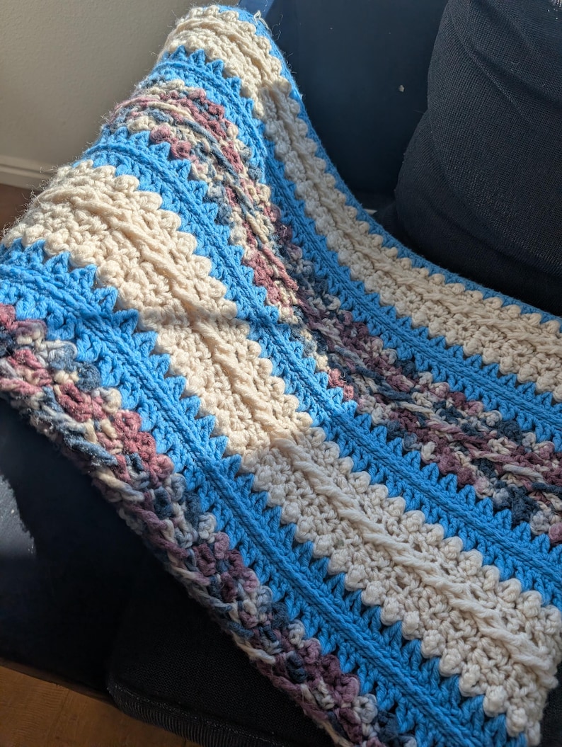 May include: A handmade crochet blanket with a textured design. The blanket features alternating stripes of light blue, cream, and muted purple. The crochet stitches create a visually appealing pattern, perfect for home decor.