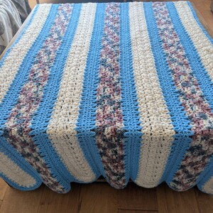 May include: A handmade crochet blanket with a striped pattern. The blanket features alternating stripes of light blue, cream, and a textured floral pattern in shades of purple and green. The edges are scalloped.