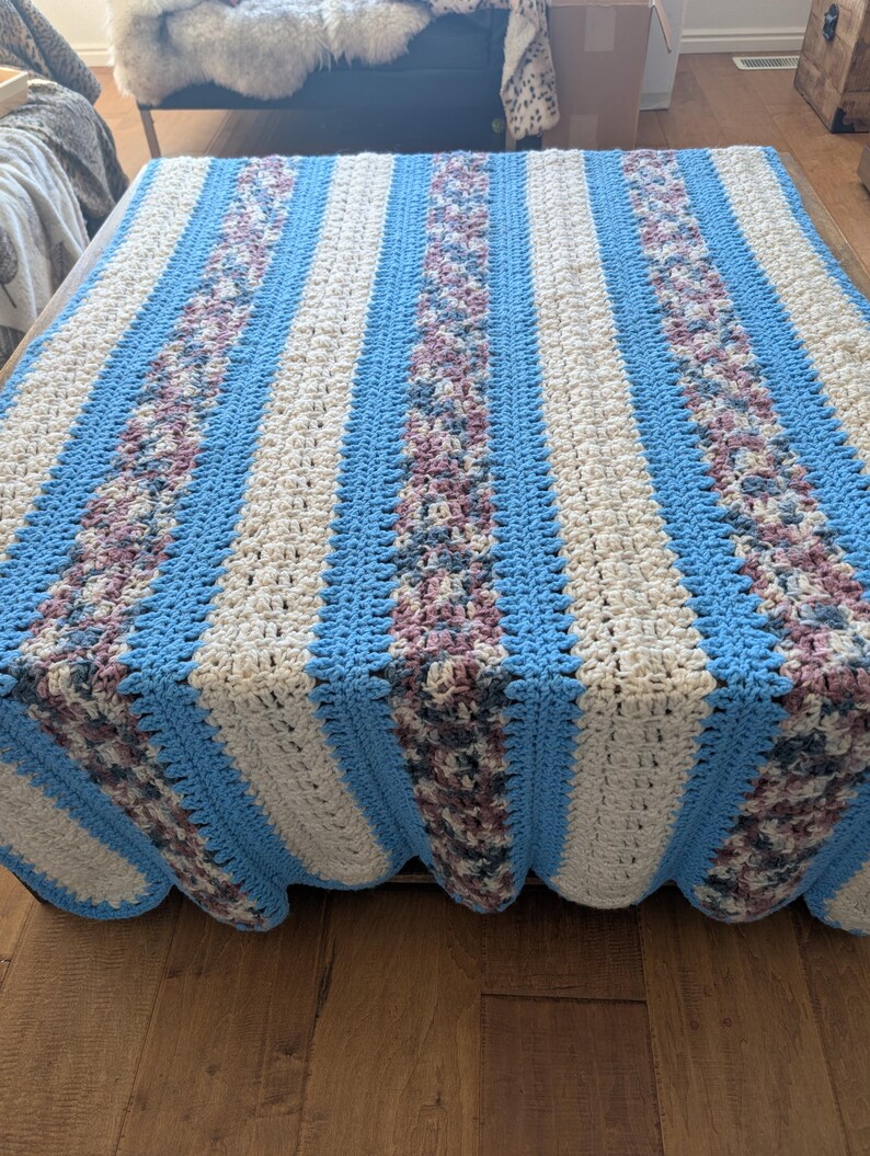 May include: A handmade crochet blanket with a striped pattern. The blanket features alternating stripes of light blue, cream, and a textured floral pattern in shades of purple and green. The edges are scalloped.