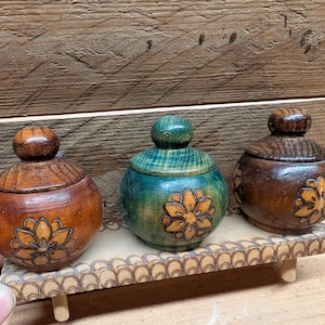 May include: Three small, round wooden lidded containers with knob handles, each featuring a wood-burned flower design. The containers are colored in light brown, teal, and dark brown, and are displayed on a rectangular wooden stand with a scalloped edge.