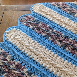 May include: A handmade crochet blanket with a scalloped edge. The blanket features a pattern of diagonal stripes in light blue, cream, and muted purple. The texture is visible, showcasing the detailed stitch work. A cozy, decorative home textile.