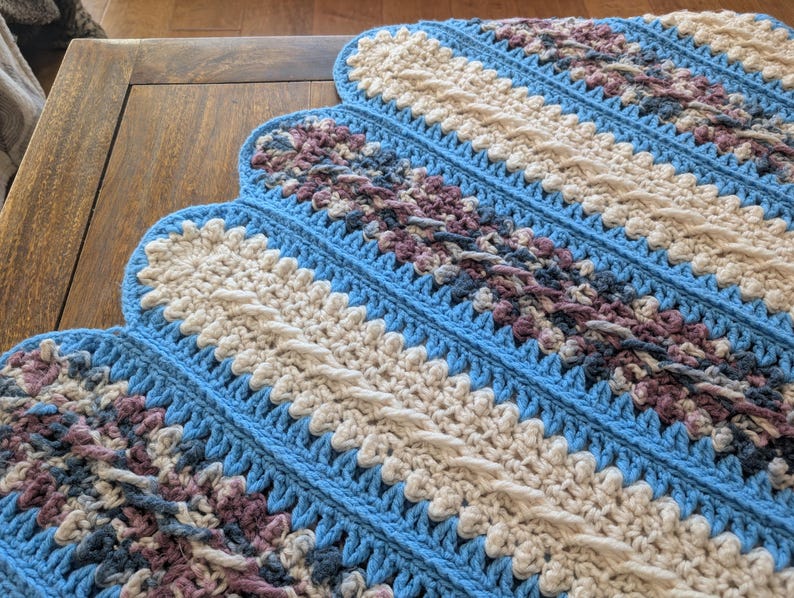 May include: A handmade crochet blanket with a scalloped edge. The blanket features a pattern of diagonal stripes in light blue, cream, and muted purple. The texture is visible, showcasing the detailed stitch work. A cozy, decorative home textile.
