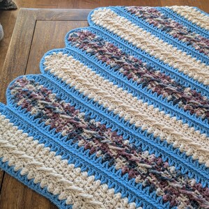 May include: A handmade crochet blanket with a scalloped edge. The blanket features alternating stripes of light blue, cream, and a patterned section with shades of purple and gray. The blanket is displayed on a wooden surface.