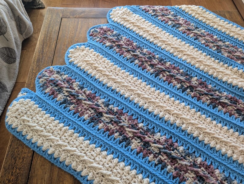 May include: A handmade crochet blanket with a scalloped edge. The blanket features alternating stripes of light blue, cream, and a patterned section with shades of purple and gray. The blanket is displayed on a wooden surface.