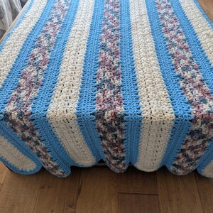 May include: A handmade crochet blanket with a striped pattern. The blanket features alternating stripes of light blue, off-white, and a textured stripe with shades of purple and gray. The blanket is draped over a wooden surface.