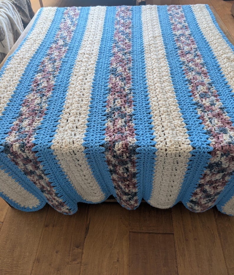 May include: A handmade crochet blanket with a striped pattern. The blanket features alternating stripes of light blue, off-white, and a textured stripe with shades of purple and gray. The blanket is draped over a wooden surface.