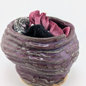 May include: A handmade, purple ceramic bowl with a textured, ridged exterior. The bowl is filled with various scrunchies in shades of black, burgundy, and gray, some adorned with pearls. The bowl sits on a light-colored base.