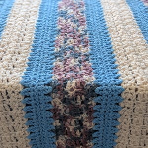 May include: A handmade crochet blanket featuring a striped pattern in light blue, cream, and a central section of muted purple and gray. The blanket has a textured appearance, with visible stitch details. The overall design is a mix of geometric and floral elements.