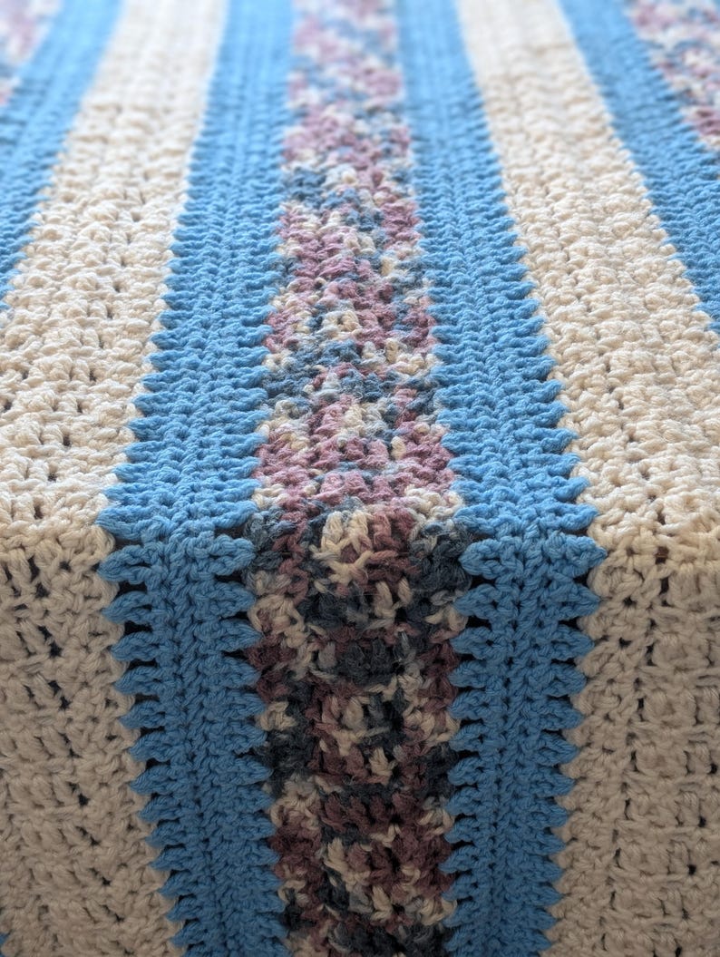 May include: A handmade crochet blanket featuring a striped pattern in light blue, cream, and a central section of muted purple and gray. The blanket has a textured appearance, with visible stitch details. The overall design is a mix of geometric and floral elements.