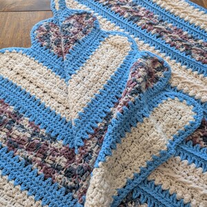May include: A handmade crochet blanket featuring a repeating pattern of blue, cream, and purple sections. The blanket has a scalloped edge and a textured design. The color scheme is a mix of cool and neutral tones.
