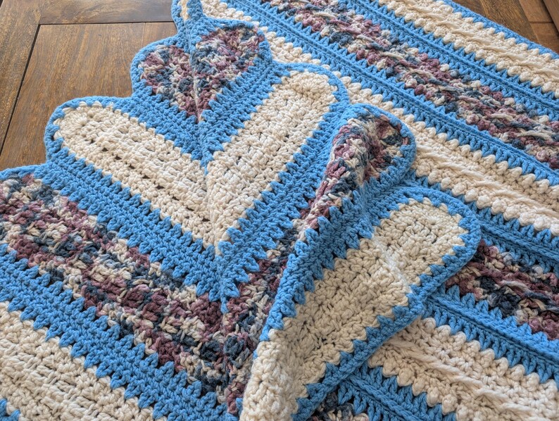 May include: A handmade crochet blanket featuring a repeating pattern of blue, cream, and purple sections. The blanket has a scalloped edge and a textured design. The color scheme is a mix of cool and neutral tones.
