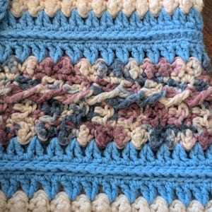 May include: Close-up of a handmade crochet item with a textured pattern. The design features alternating bands of light blue, cream, and a mix of muted pink and blue yarns. The stitches create a raised, tactile surface.