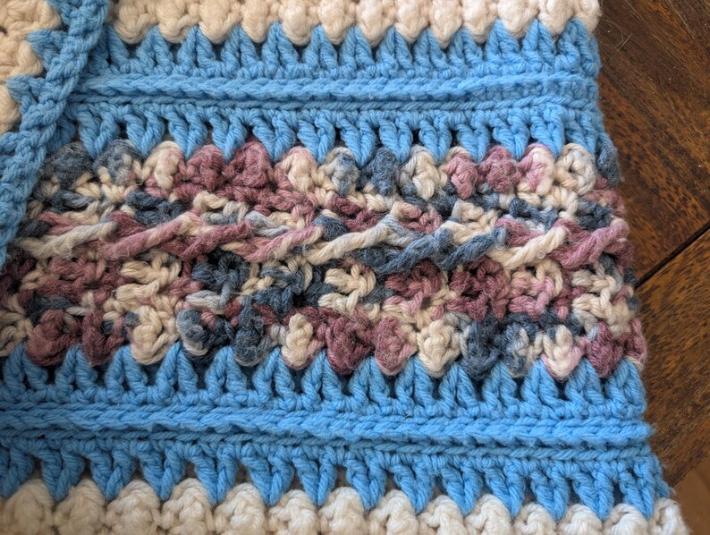 May include: Close-up of a handmade crochet item with a textured pattern. The design features alternating bands of light blue, cream, and a mix of muted pink and blue yarns. The stitches create a raised, tactile surface.